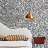 A modernist bed, hanging lamp and chair stand in front of a wall papered in an abstract linear print in black and white.