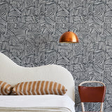 A modernist bed, hanging lamp and chair stand in front of a wall papered in an abstract linear print in white and charcoal.