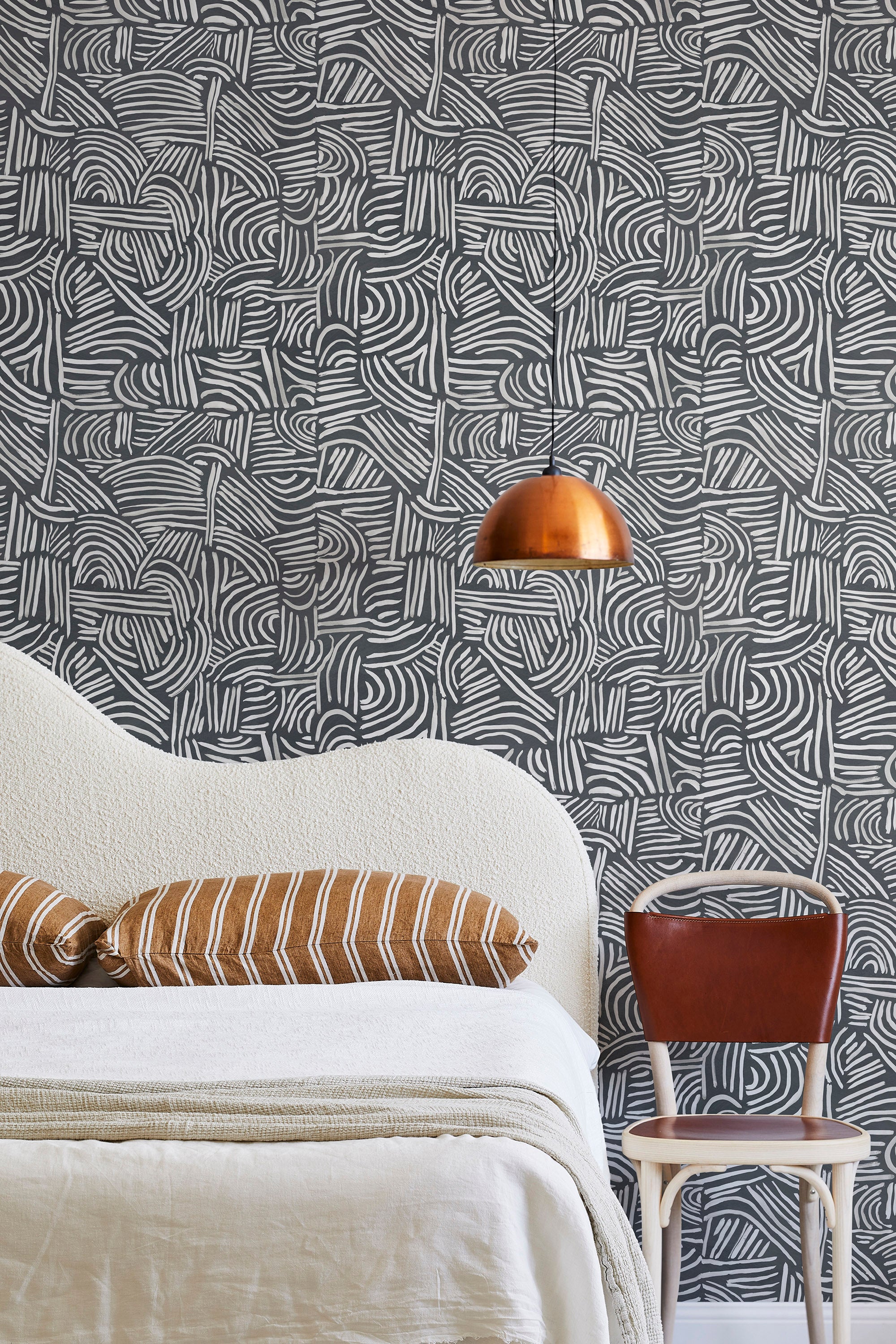 A modernist bed, hanging lamp and chair stand in front of a wall papered in an abstract linear print in white and charcoal.