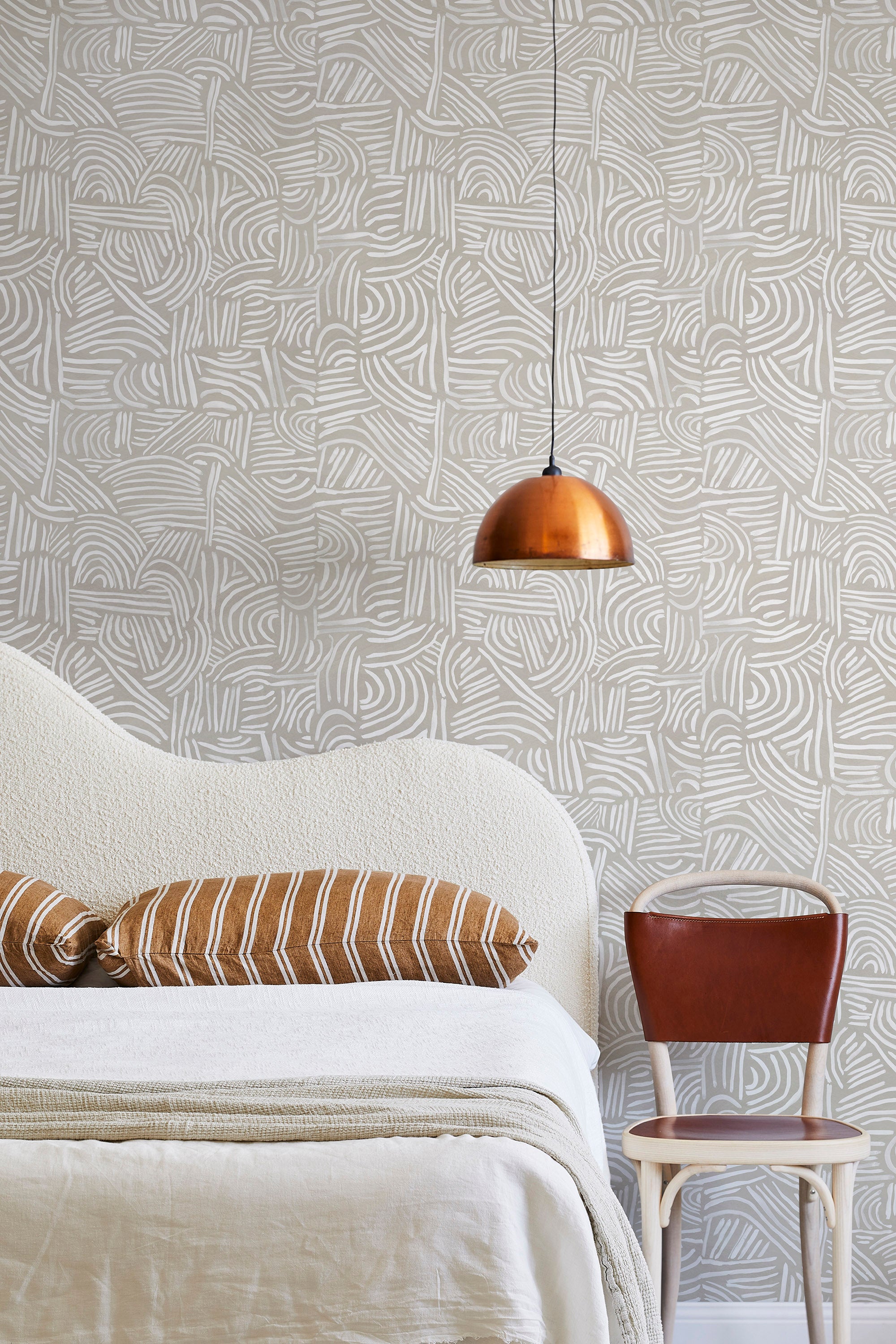 A modernist bed, hanging lamp and chair stand in front of a wall papered in an abstract linear print in white and cream.