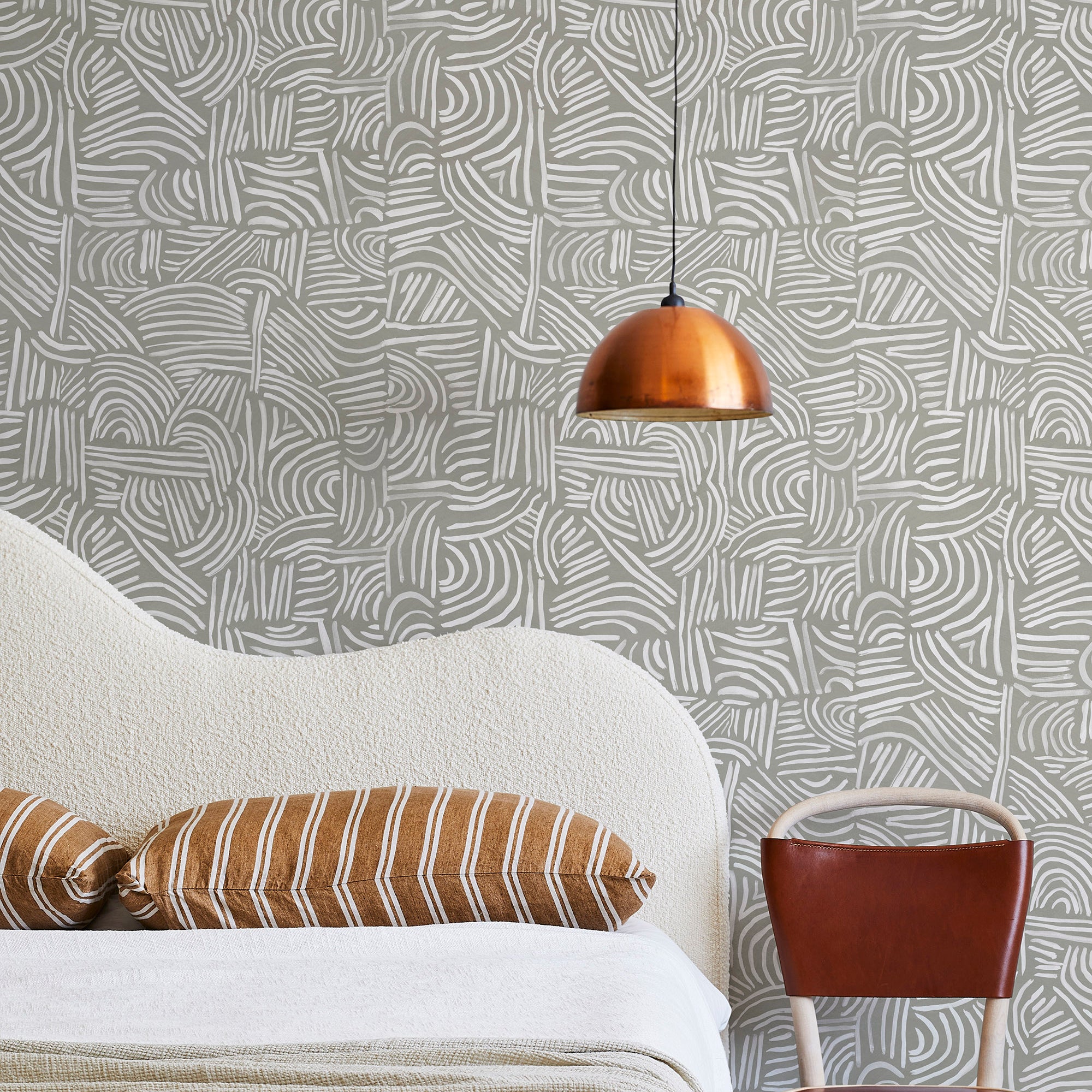 A modernist bed, hanging lamp and chair stand in front of a wall papered in an abstract linear print in white and sage.