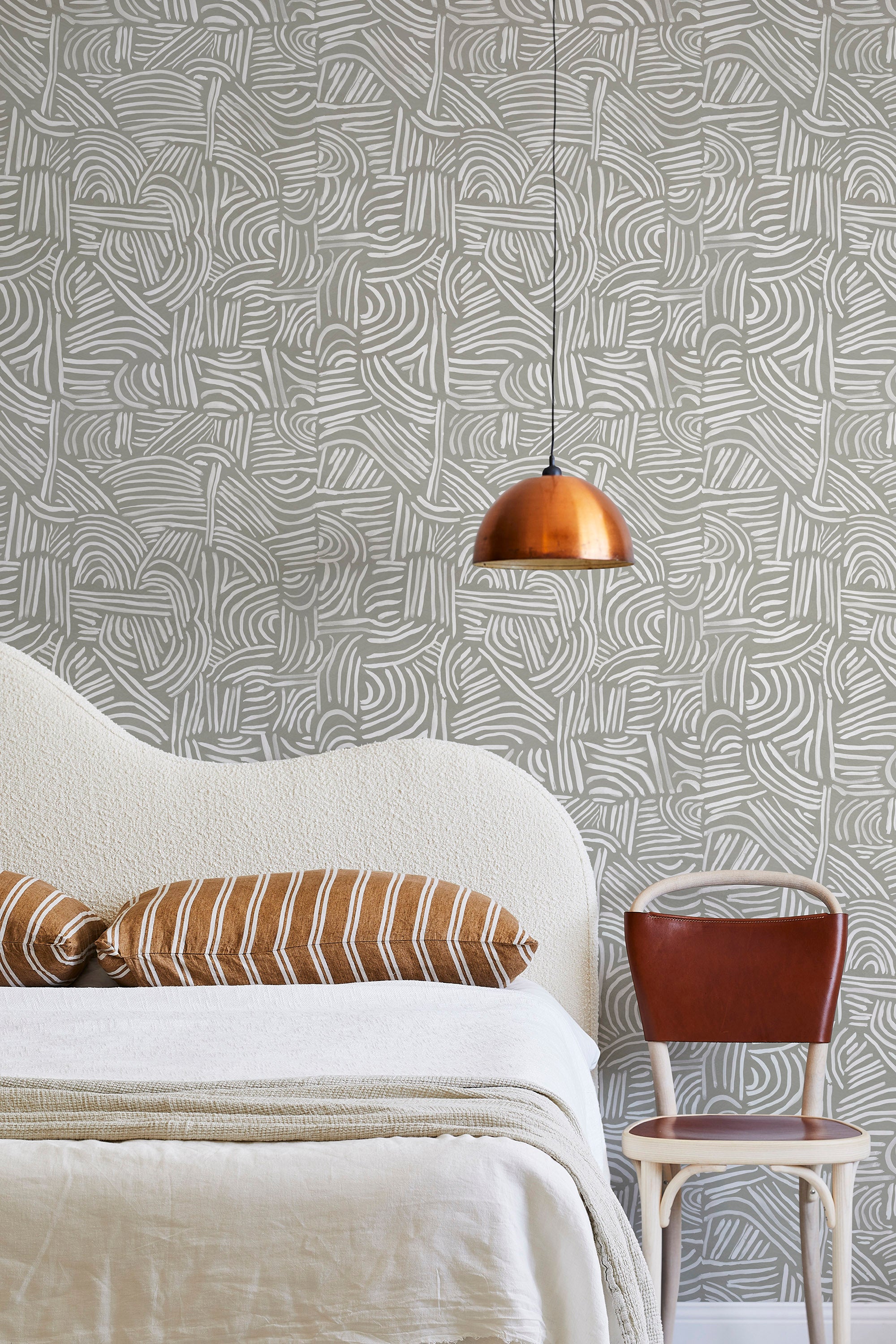 A modernist bed, hanging lamp and chair stand in front of a wall papered in an abstract linear print in white and sage.