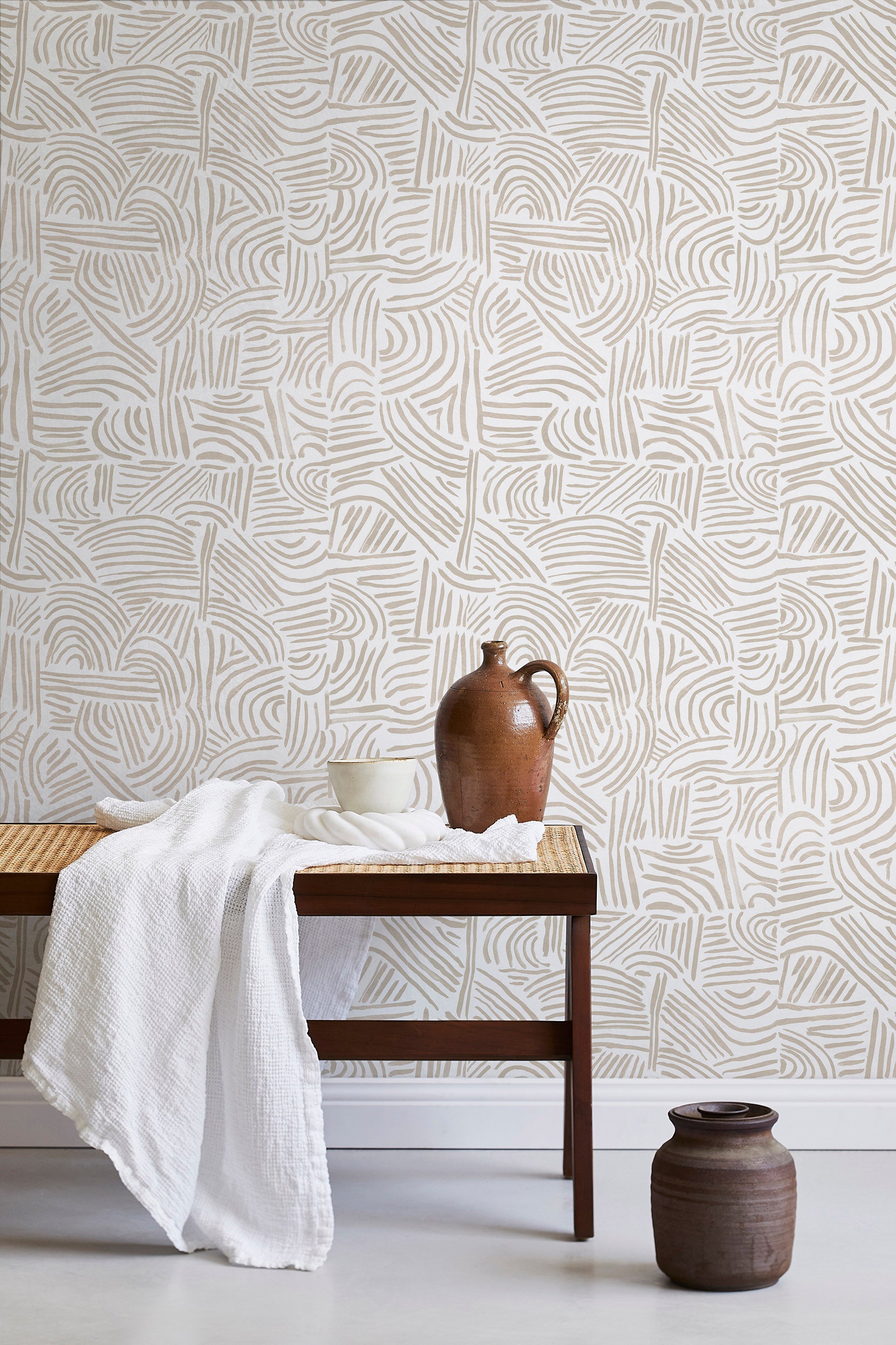 A bench with a towel and clay vases stands in front of a wall papered in an abstract linear print in cream and white.