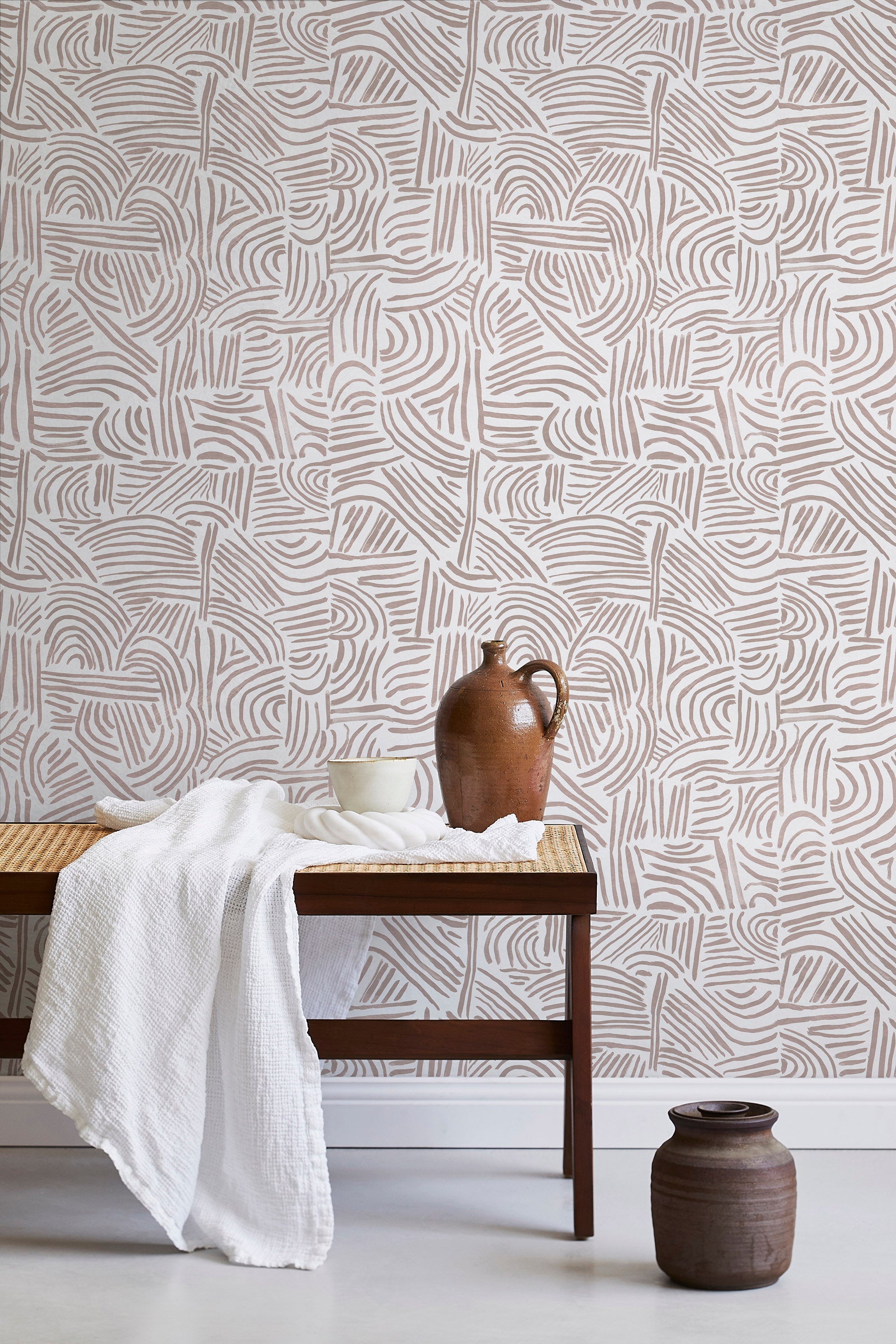 A bench with a towel and clay vases stands in front of a wall papered in an abstract linear print in blush and white.