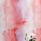 A vase of flowers stands in front of a wall papered in an abstract curvilinear pattern in mottled pink, red and purple.