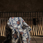 Chair draped in a woven performance fabric in a botanical branch and stripe design in beige, tan and red on a navy blue field against a metal railing and stone wall. 