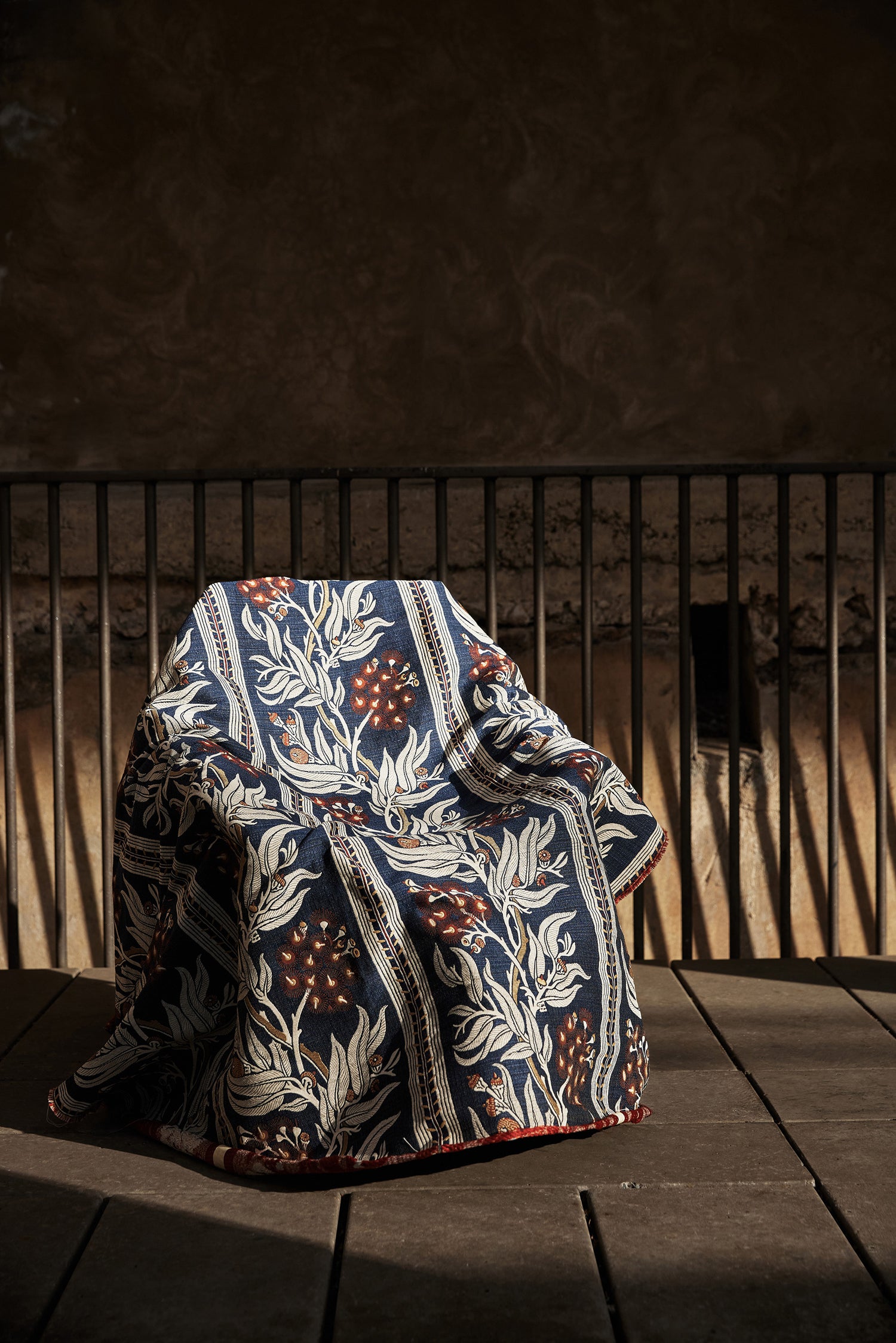 Chair draped in a woven performance fabric in a botanical branch and stripe design in beige, tan and red on a navy blue field against a metal railing and stone wall. 