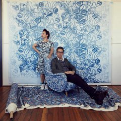 Portrait of a couple in front of a floral backdrop: a man sitting on a patterned chair with his legs spread out, and a woman standing next to the chair.