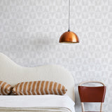 A modernist bed, hanging lamp and chair stand in front of a wall papered in a curvilinear grid print in white and light gray.