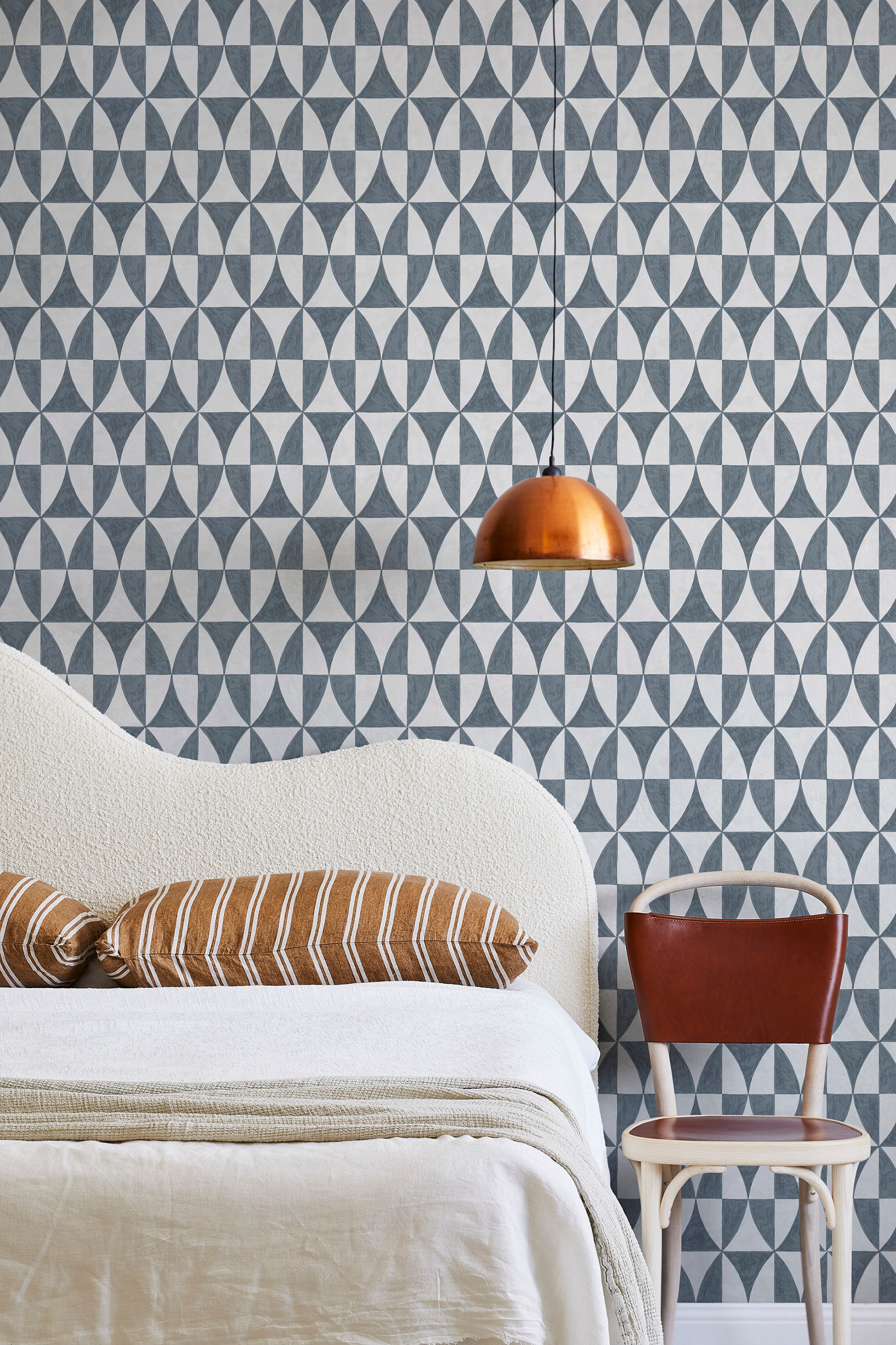 A modernist bed, hanging lamp and chair stand in front of a wall papered in a curvilinear grid print in navy and white.