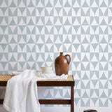 A bench with a towel and clay vases stands in front of a wall papered in a curvilinear grid print in light blue and white.