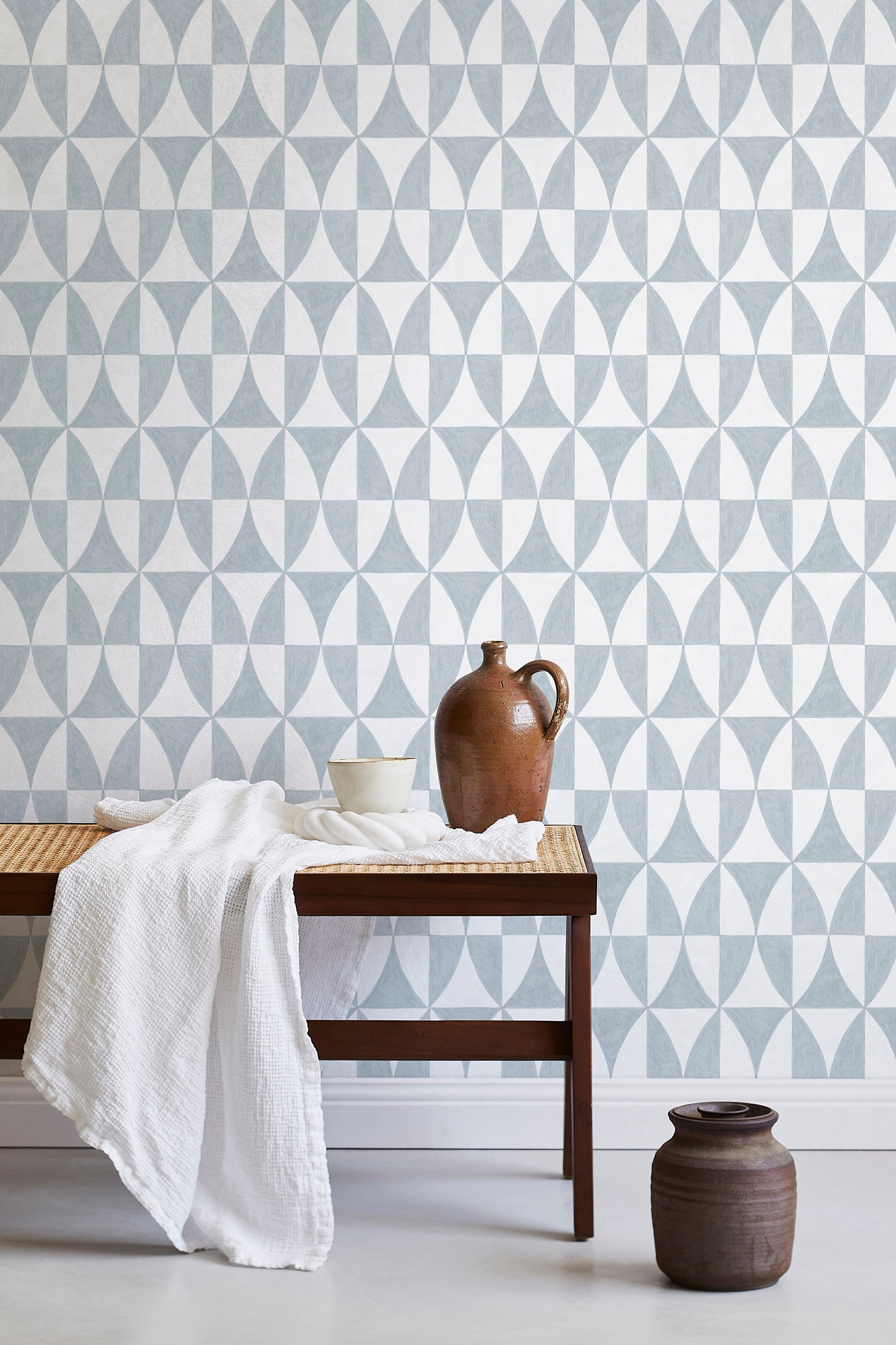 A bench with a towel and clay vases stands in front of a wall papered in a curvilinear grid print in light blue and white.