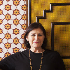 Close-up of an older woman with a brunette bob standing in front of a wall with colorful wallpaper and wood trim.