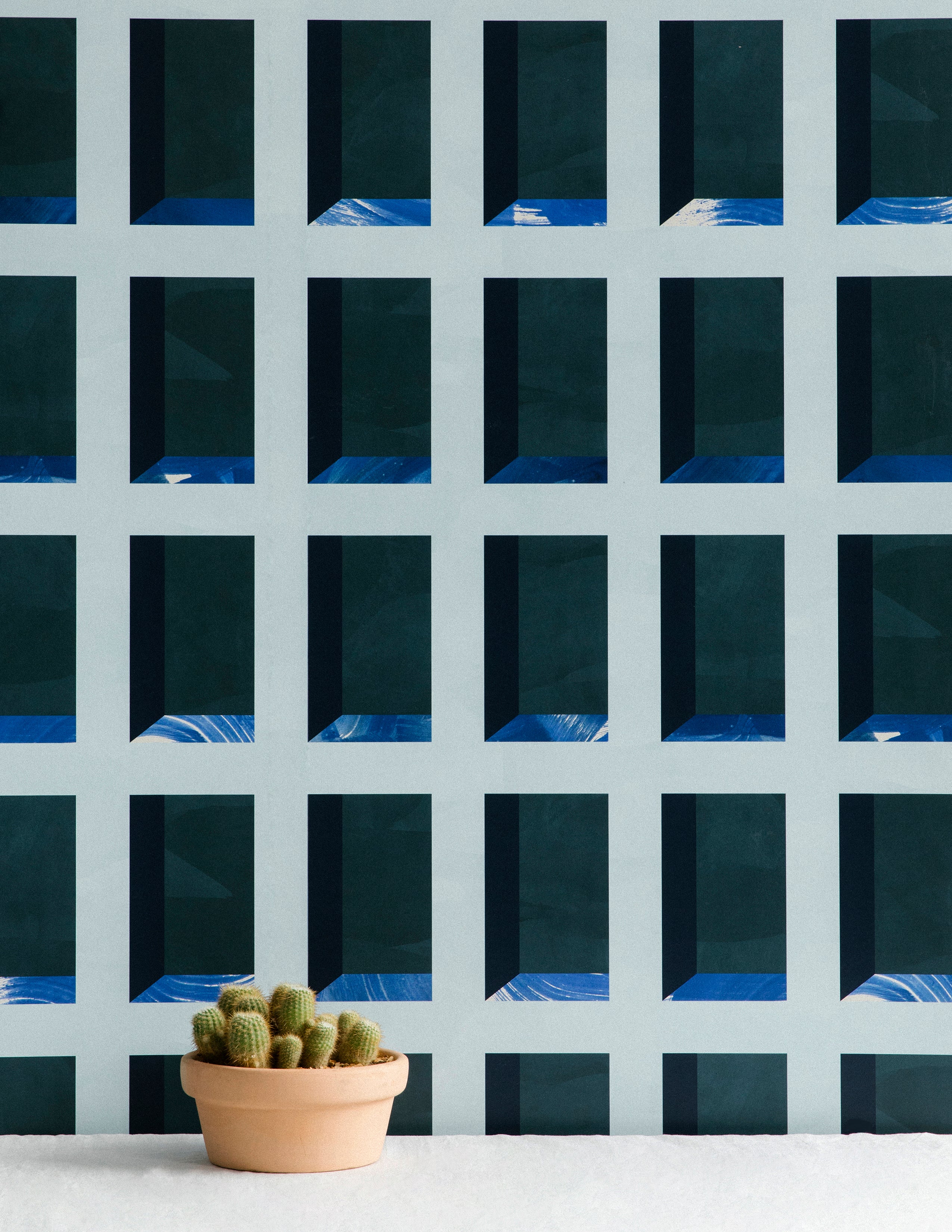 A potted cactus stands in front of a wall papered in a dimensional grid print in shades of blue, navy and turquoise.