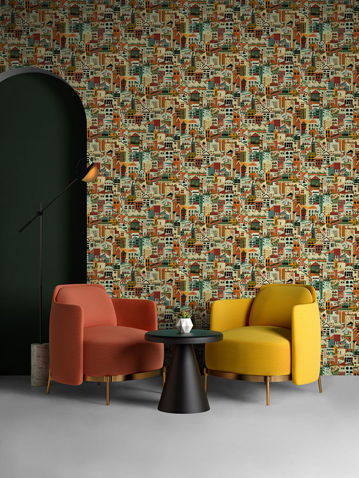 A pair of armchairs in front of a wall papered in a repeating building print in shades of blue, orange, red and green. 