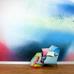 A sofa and throw pillows stands in front of a wall covered in an ombré paint splatter print in blue, navy, red and white.