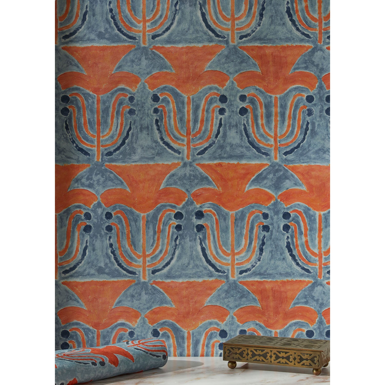 A small bronze filigree box and folded fabric swatch in front of a wallpaper with stamped painted floral motif in red and cobalt on a medium blue background, hover