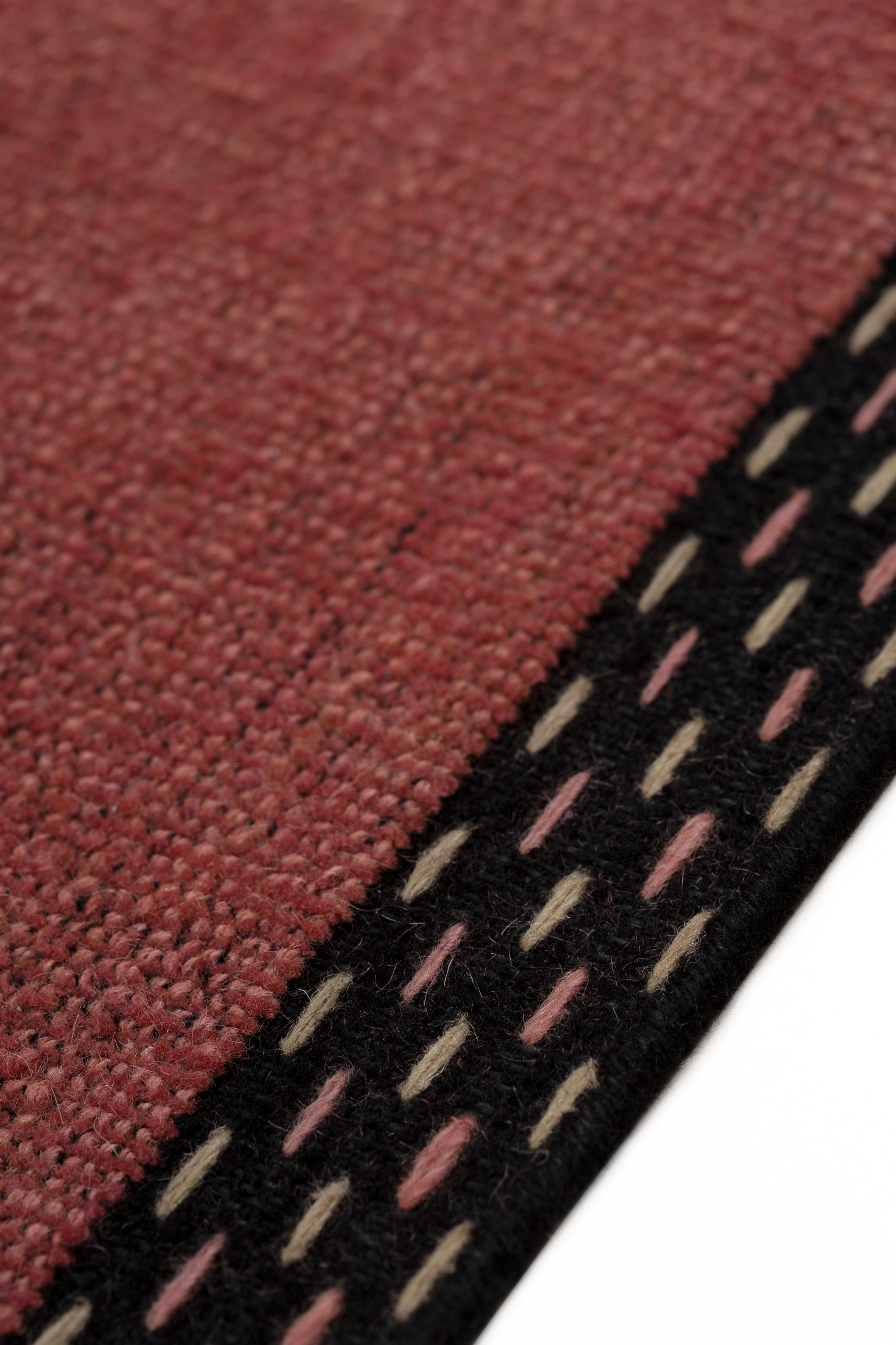 Detail of the Plain Stitched Border Rug in Carmine, a solid red rug with a black border with black border with pink and taupe dashes. 