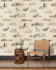 A woven chair and end table in front of a wall papered in a pattern of Japanese soldiers and horses carrying spears and flags.