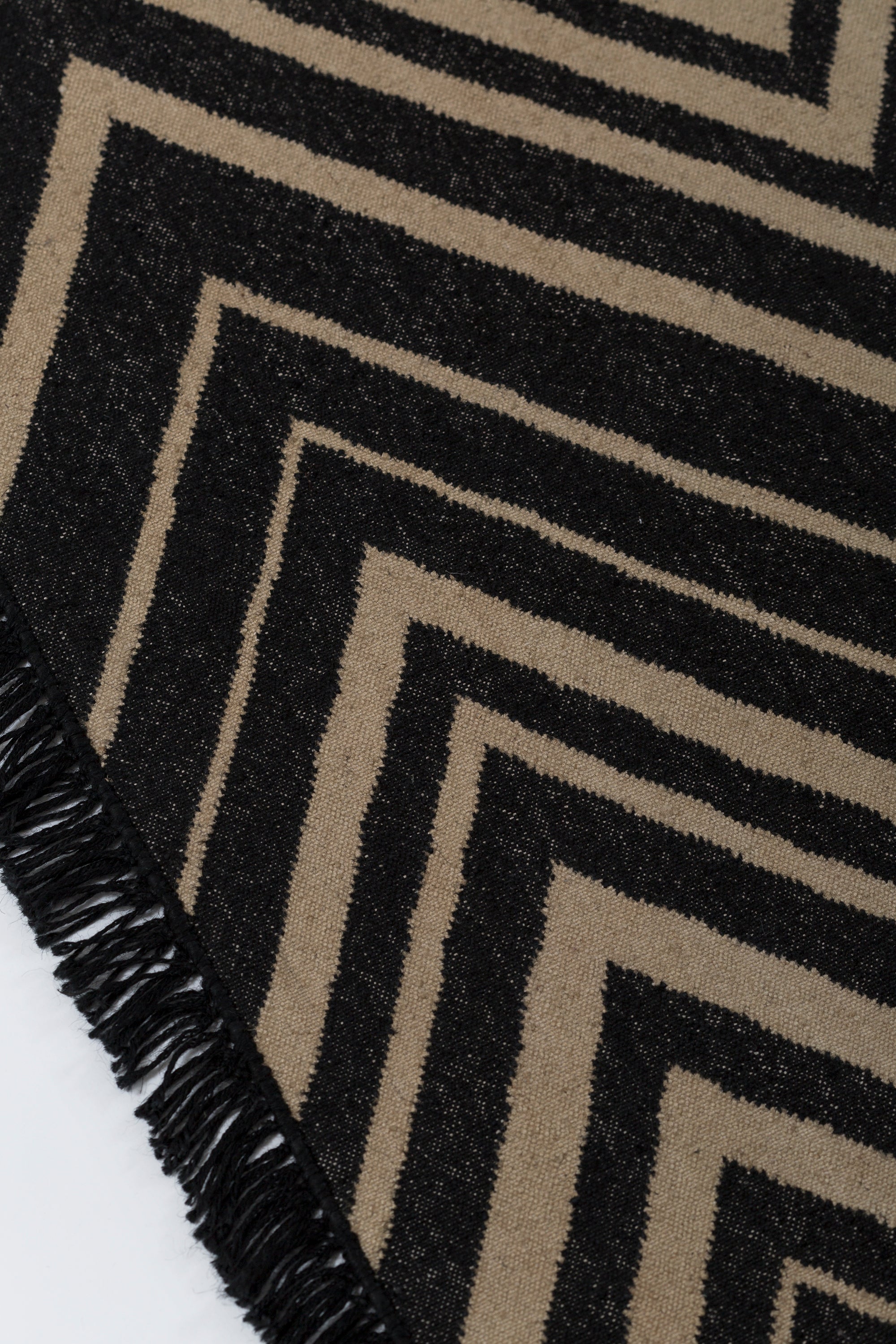 Detail of the Vertical Zig-Zag Rug in Onyx-Sandstone, a zig zag pattern in mixed widths in black and taupe with a fringed edge. 