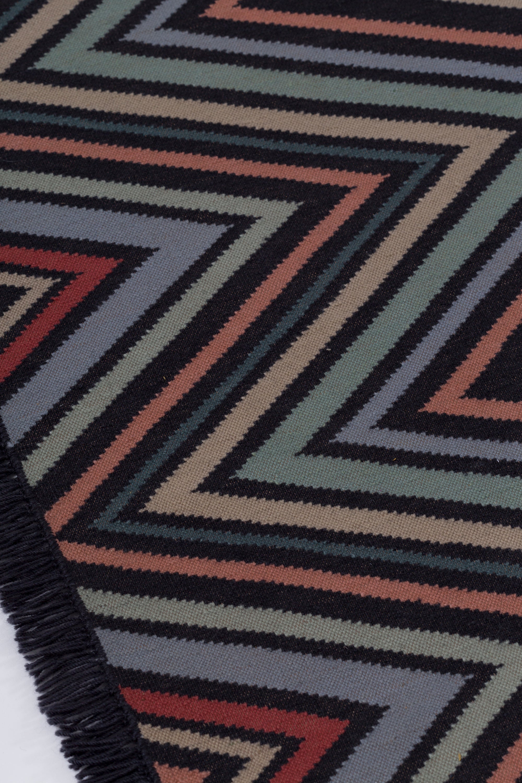 Detail of the To The Point Rug in Hessonite-Garnet-Slate, a zig zag pattern in red, blue coral with black, with a black fringed edge. 