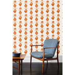 A wooden chair and coffee table in front of a wall papered in a large-scale pattern of orange watercolored diamonds, circles and lines on a cream background. 