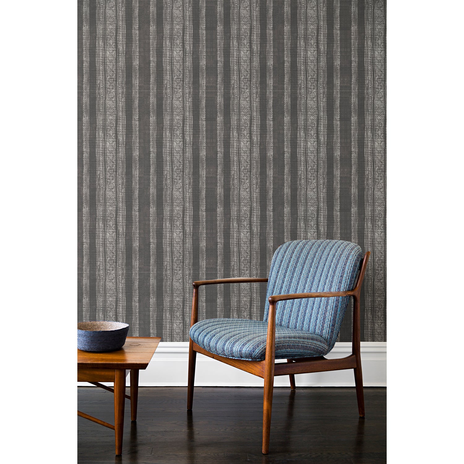 An armchair and coffee table in front of a wall papered in a striped pattern of intricate white lines over a washed gray background.