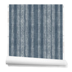 A partially unrolled roll of wallpaper in a striped pattern of intricate white lines over a washed denim background.