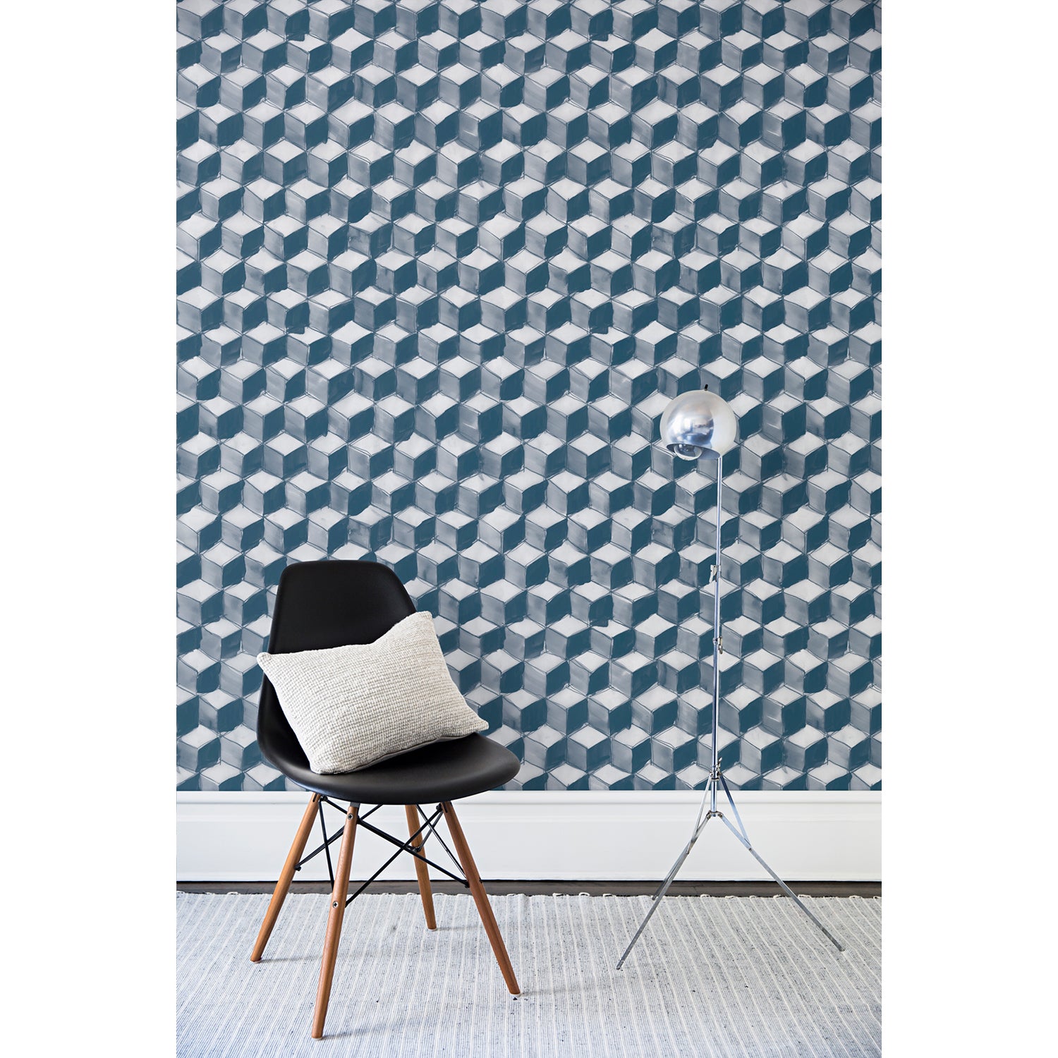 A chair and floor lamp in front of a wall papered in a hand-painted tumbling block pattern in shades of blue on a white background.