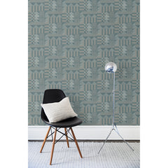 A wall papered in layered geometric shapes in tan, navy and gray with a black chair and silver floor lamp in front of it.