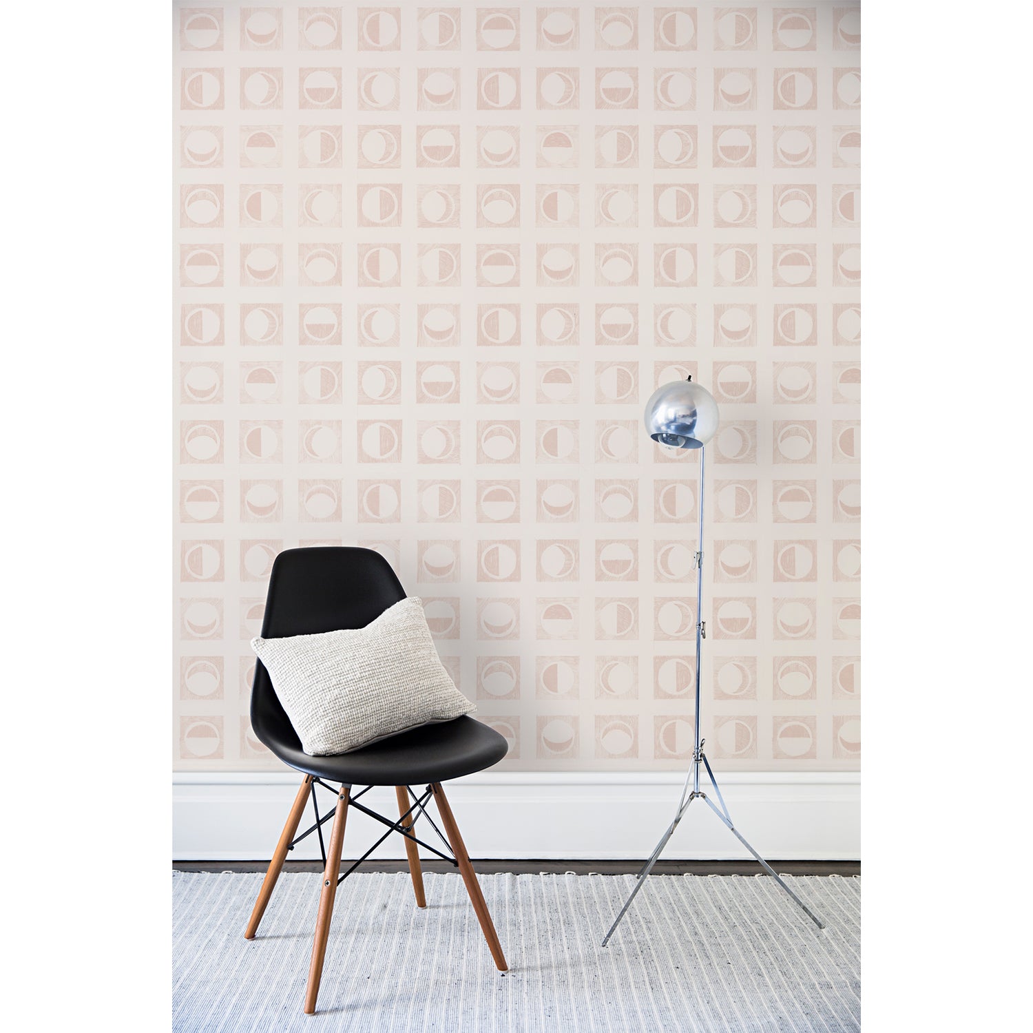 A chair and floor lamp in front of a wall papered in rows of block-printed light pink moons in various phases on a tan background.