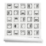 Partially unrolled wallpaper roll with rows of block-printed moons in various phases. Moons are black on a white background.