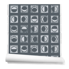 Partially unrolled wallpaper roll with rows of block-printed moons in various phases. Moons are white on a charcoal background.