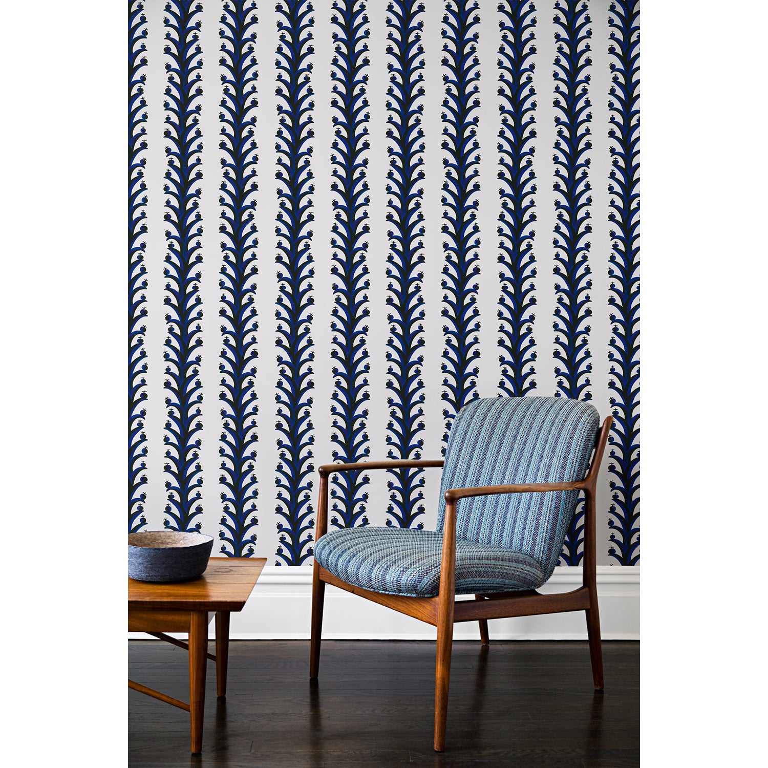 An armchair and coffee table in front of a wall papered in repeating horizontal stripes of black and blue branches and fruit on a white background.