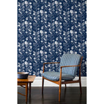 An armchair and coffee table in front of a wall papered in repeating rows of colorful leaf prints in shades of blue and white on a navy background.